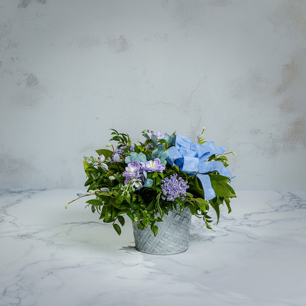 Fresh salal and green huckleberry with faux lilac delphinium flowers, faux purple verbena, faux pussy willows, and a periwinkle bow in a metal white patterned pot