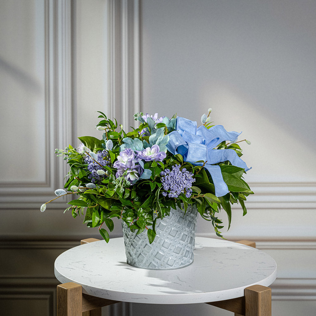 Fresh salal and green huckleberry with faux lilac delphinium flowers, faux purple verbena, faux pussy willows, and a periwinkle bow in a metal white patterned pot