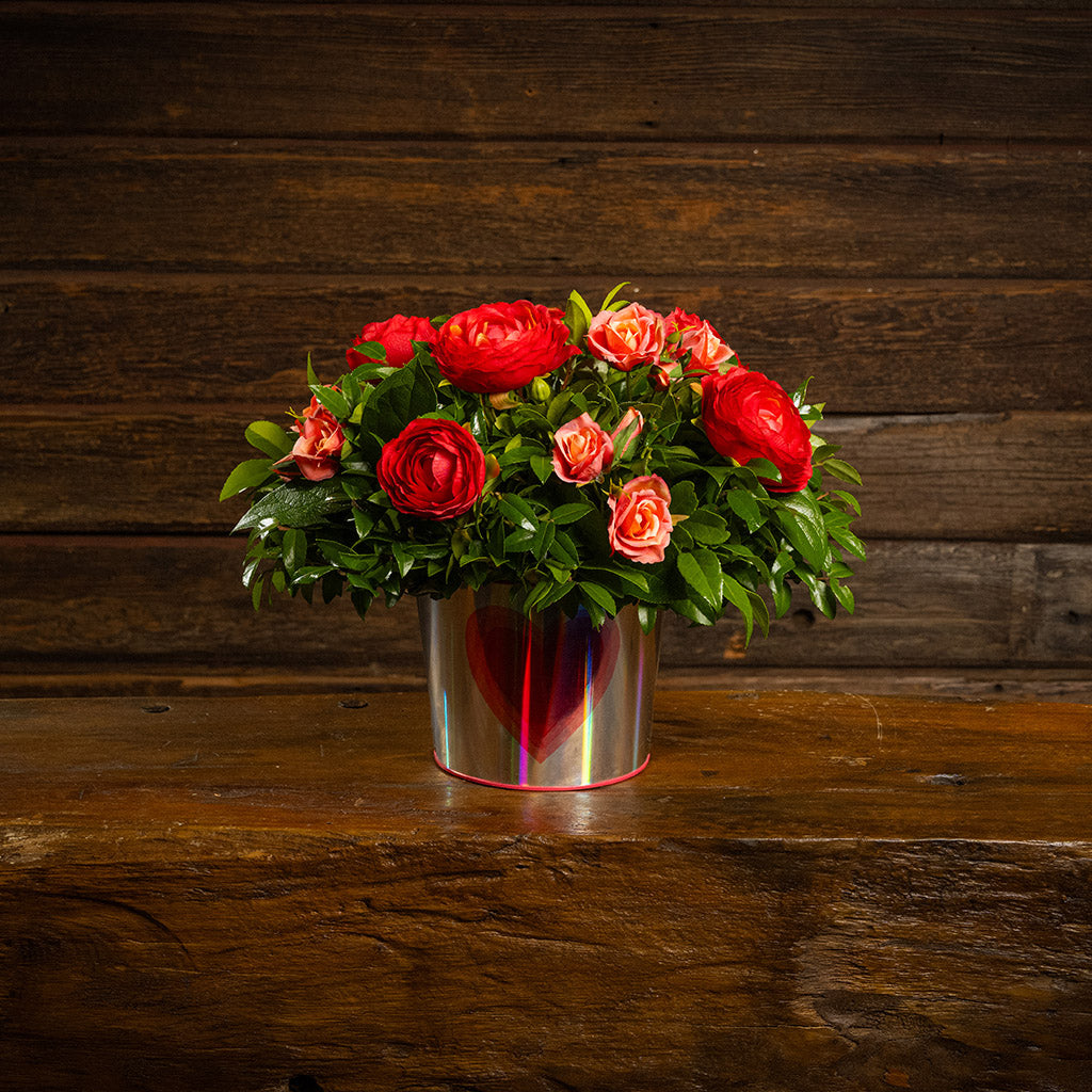 Salal and huckleberry with faux red ranunculus flowers and faux peach roses in a silver metal container adorned with hearts