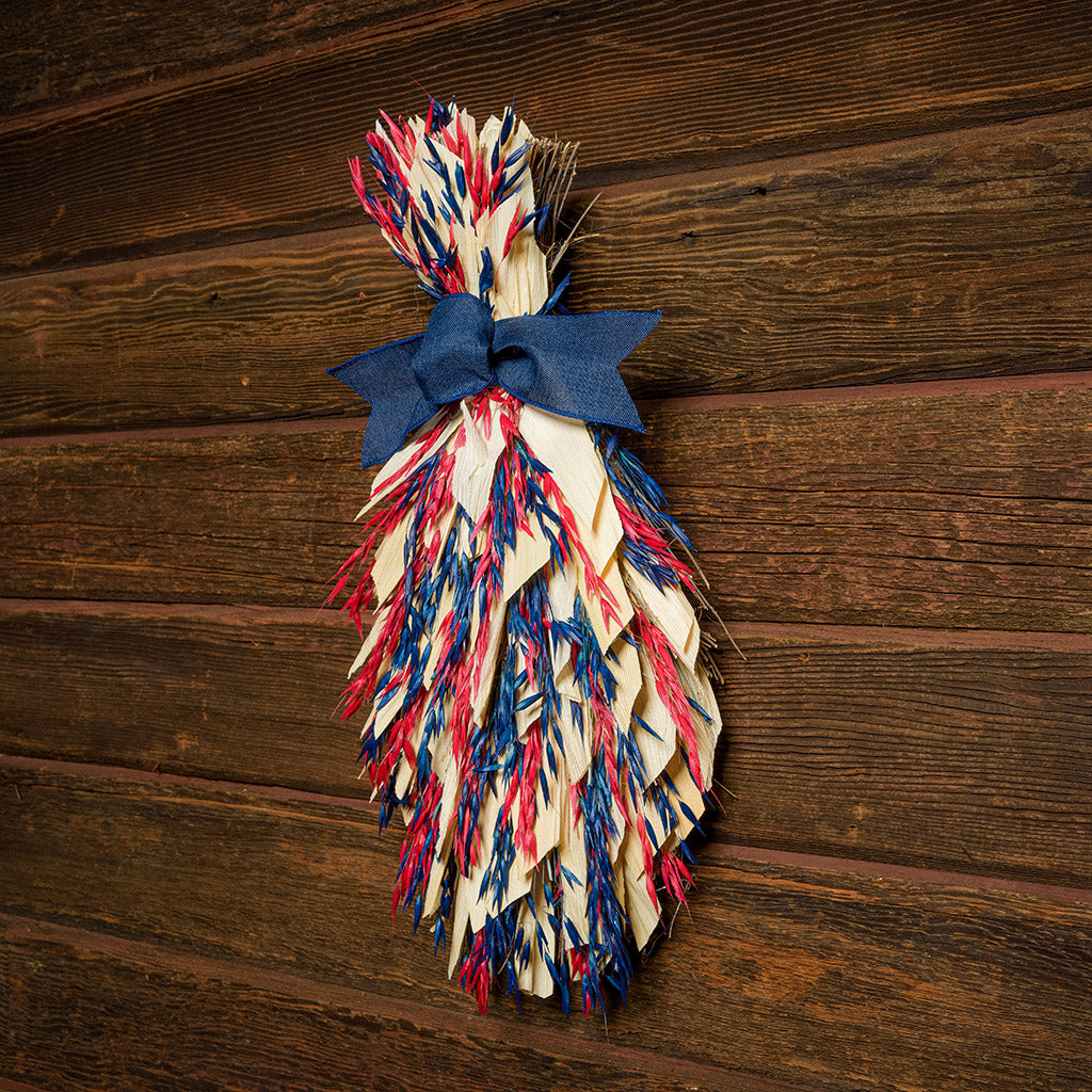 Floral bouquet of red and blue oats and natural corn husks and tied together with a navy-blue canvas bow on a dark wood background.