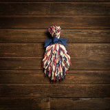 Floral bouquet of red and blue oats and natural corn husks and tied together with a navy-blue canvas bow on a dark wood background.