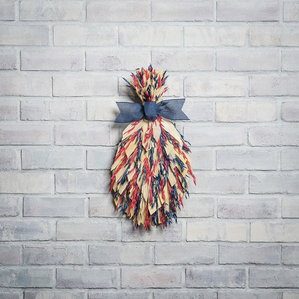 Floral bouquet of red and blue oats and natural corn husks and tied together with a navy-blue canvas bow on a white brick background.