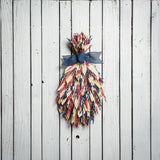 Floral bouquet of red and blue oats and natural corn husks and tied together with a navy-blue canvas bow on a white wood fence background.