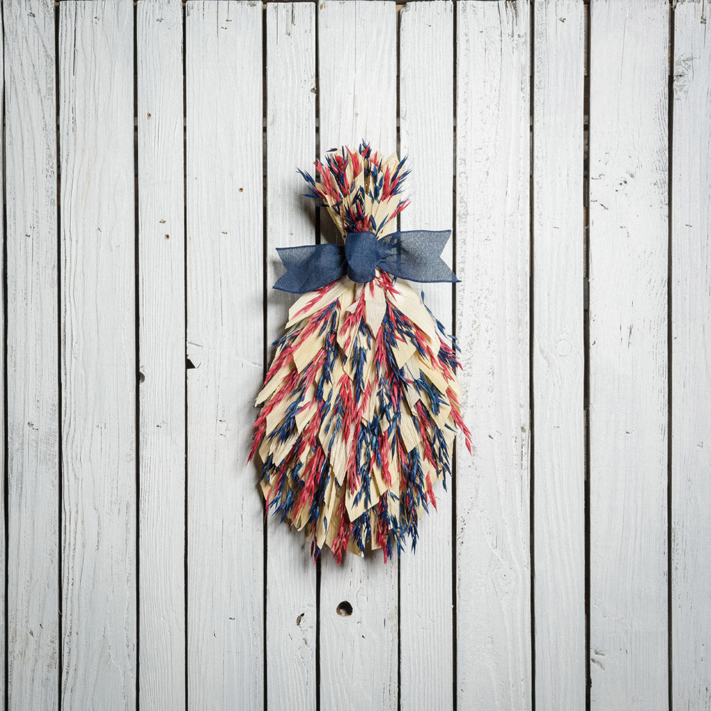 Floral bouquet of red and blue oats and natural corn husks and tied together with a navy-blue canvas bow on a white wood fence background.