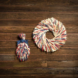 Floral bouquet and 22" wreath of red and blue oats and natural corn husks and tied together with a navy-blue canvas bow on a dark wood background.