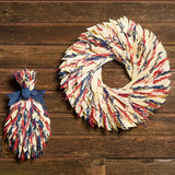 Floral bouquet and 22" wreath of red and blue oats and natural corn husks and tied together with a navy-blue canvas bow on a dark wood background.