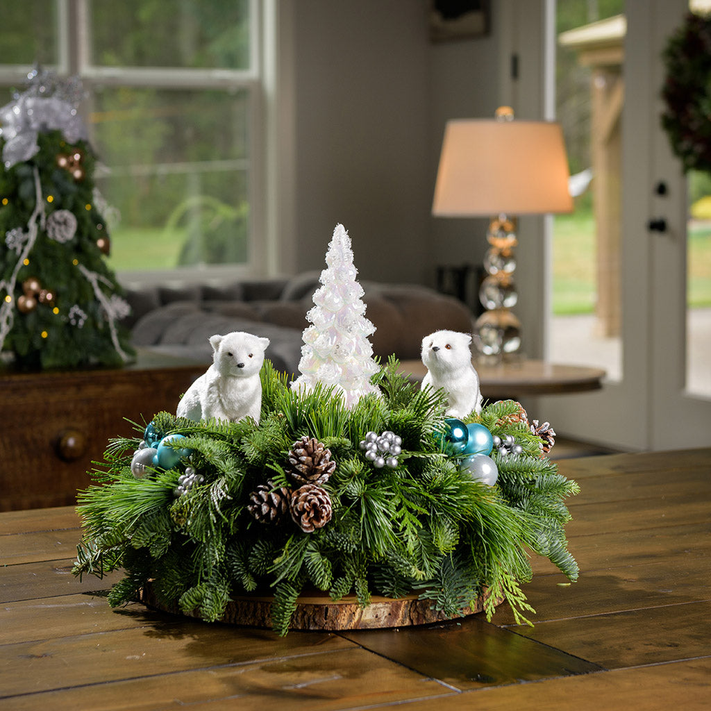 Centerpiece made of noble fir, white pine, incense cedar, aqua-blue ball clusters, white-tipped pinecones, silver berry clusters, 2 polar bears, and a shiny light-up glass tree on a wooden table against a wood wall