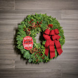 A holiday wreath of noble fir and white pine with faux red berries, 5 Australian cones, a keepsake red enamel “Believe in the Magic of Christmas” sign, and a red brushed-linen bow on a wood background.
