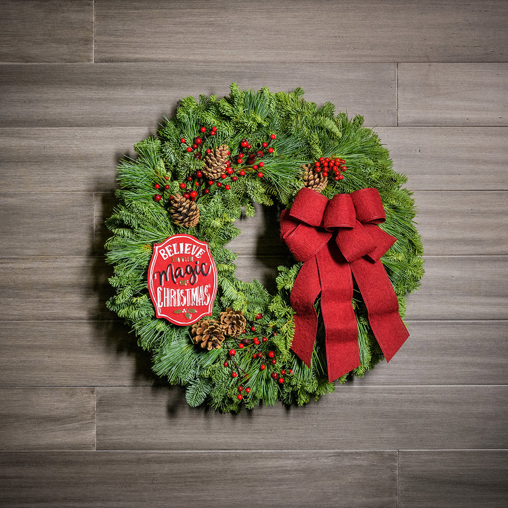 A holiday wreath of noble fir and white pine with faux red berries, 5 Australian cones, a keepsake red enamel “Believe in the Magic of Christmas” sign, and a red brushed-linen bow on a wood background.