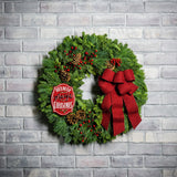 A holiday wreath of noble fir and white pine with faux red berries, 5 Australian cones, a keepsake red enamel “Believe in the Magic of Christmas” sign, and a red brushed-linen bow on a white brick background.