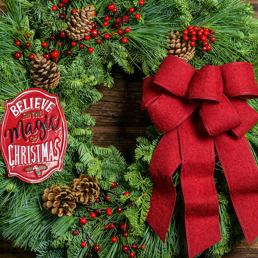 A holiday wreath of noble fir and white pine with faux red berries, 5 Australian cones, a keepsake red enamel “Believe in the Magic of Christmas” sign, and a red brushed-linen bow on a wood background.