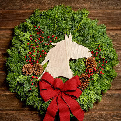 Christmas wreath made of noble fir and cedar with a wooden horse head cutout, 4 pine cones, red berry clusters, and a red brushed linen bow on a dark wood background.