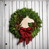 Christmas wreath made of noble fir and cedar with a wooden horse head cutout, 4 pine cones, red berry clusters, and a red brushed linen bow on a white wood background.