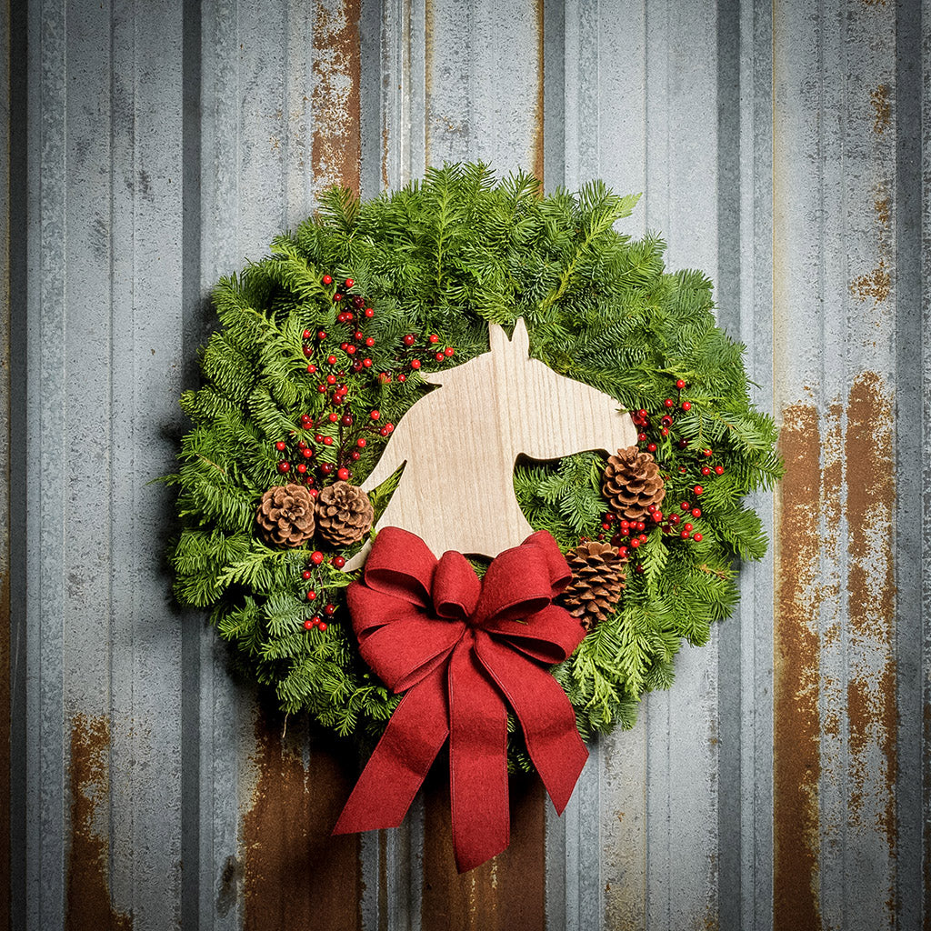 Christmas wreath made of noble fir and cedar with a wooden horse head cutout, 4 pine cones, red berry clusters, and a red brushed linen bow on a rustic metal background.