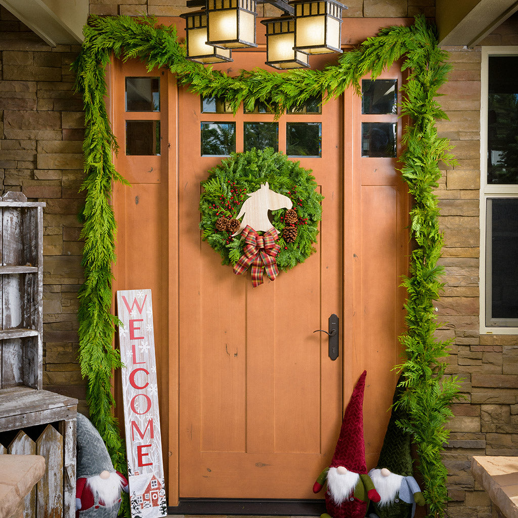 White-washed with a red barn in a wintery holiday snow scene is accented with trees and falling snowflakes with "Welcome" written in large red letters paired with the Best In Show wreath, plush gnomes, and garland on a front porch.