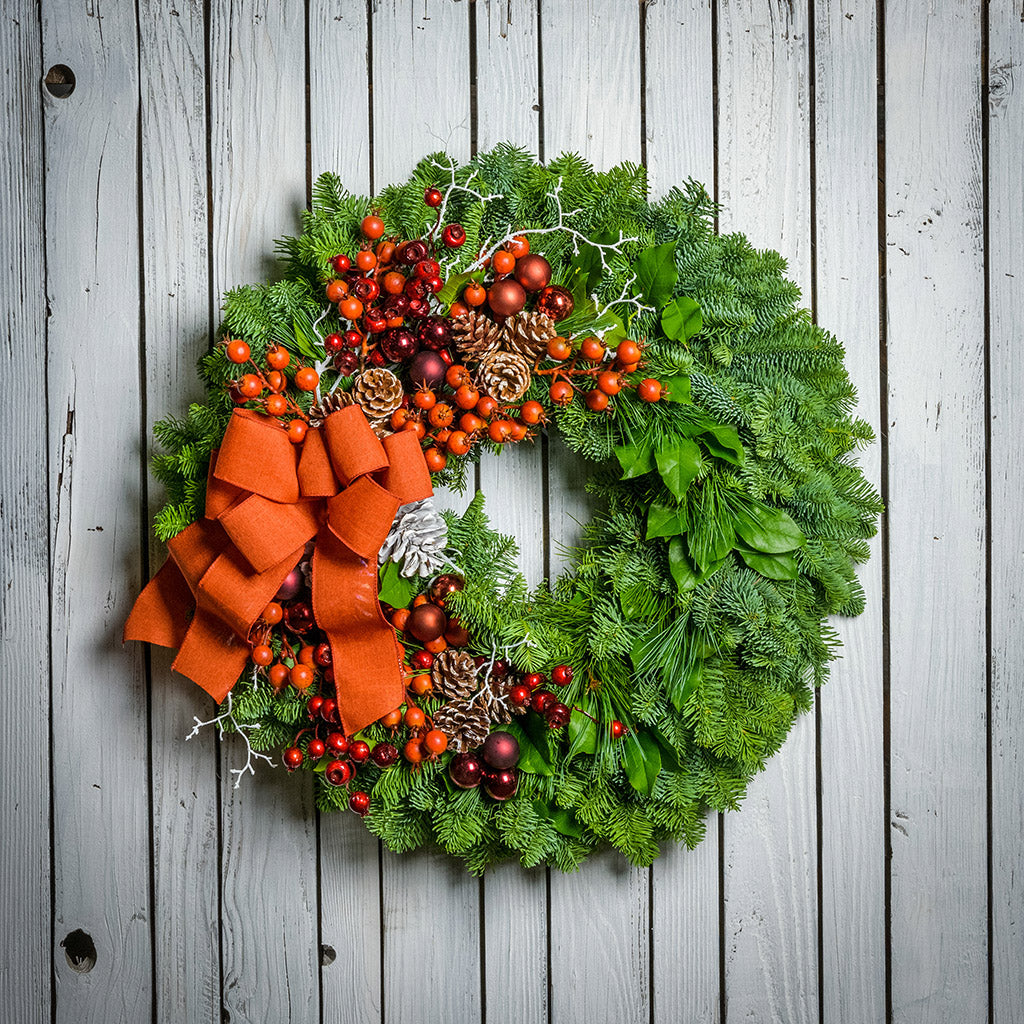 Holiday wreath made of noble fir, pine, and salal leaves with orange and metallic faux berries, 3 burgundy and 3 copper-colored ball clusters, white branches, 11 white-tipped pine cones, and an orange bow hanging on a white fence