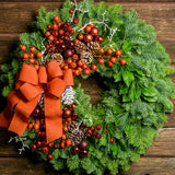 Holiday wreath made of noble fir, pine, and salal leaves with orange and metallic faux berries, 3 burgundy and 3 copper-colored ball clusters, white branches, 11 white-tipped pine cones, and an orange bow
