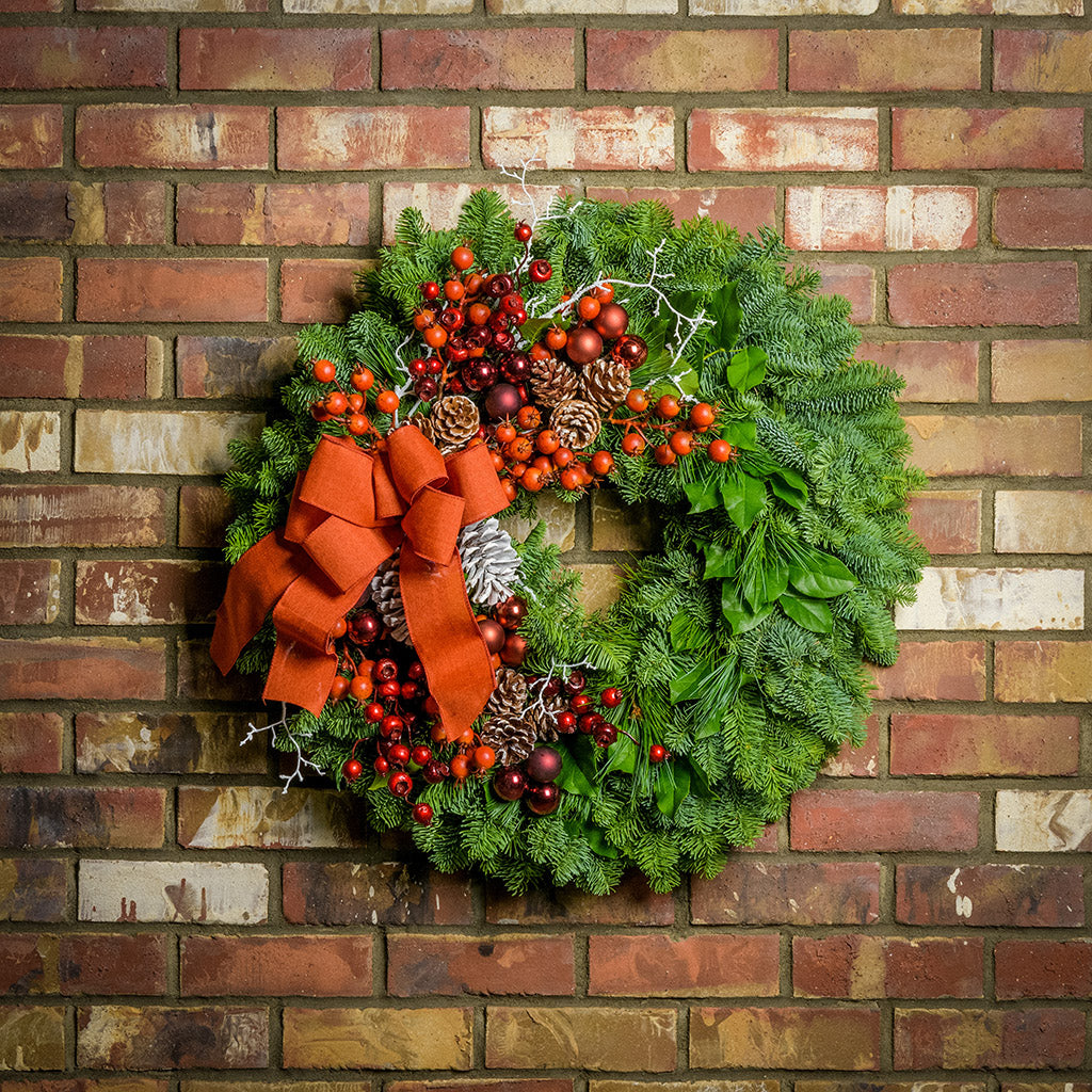 Holiday wreath made of noble fir, pine, and salal leaves with orange and metallic faux berries, 3 burgundy and 3 copper-colored ball clusters, white branches, 11 white-tipped pine cones, and an orange bow hanging on a brick wall