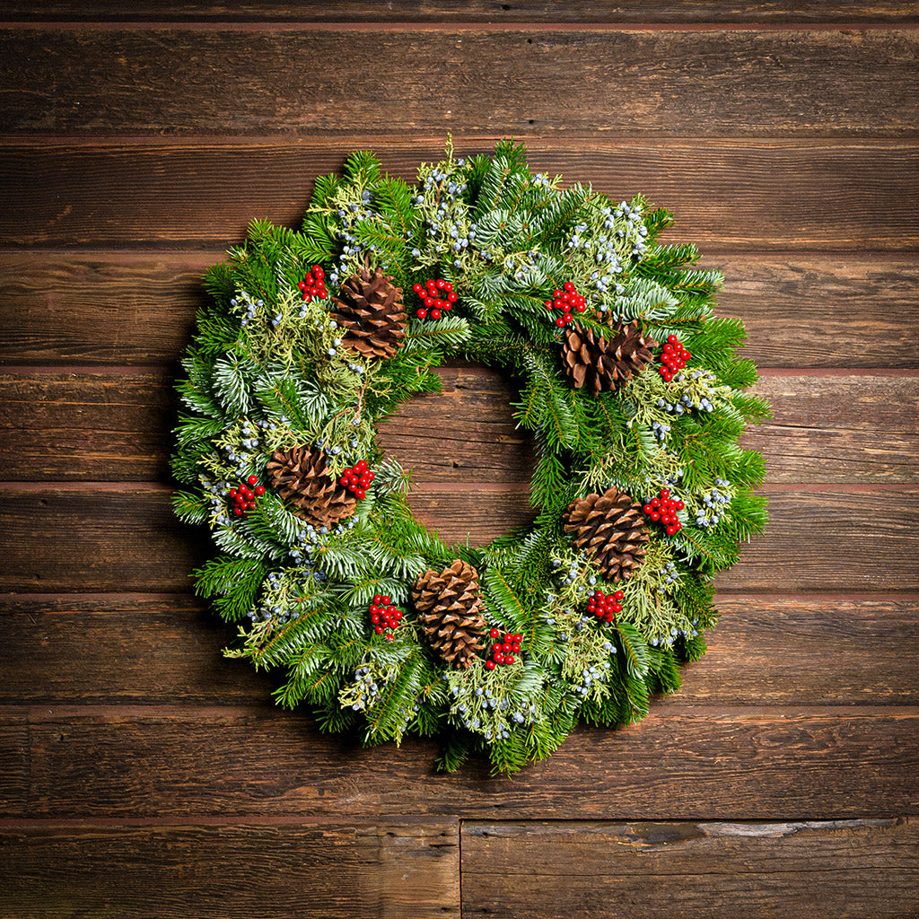 A wreath made of Nordmann fir and juniper with ponderosa pine cones and red berries with a dark wooden background.