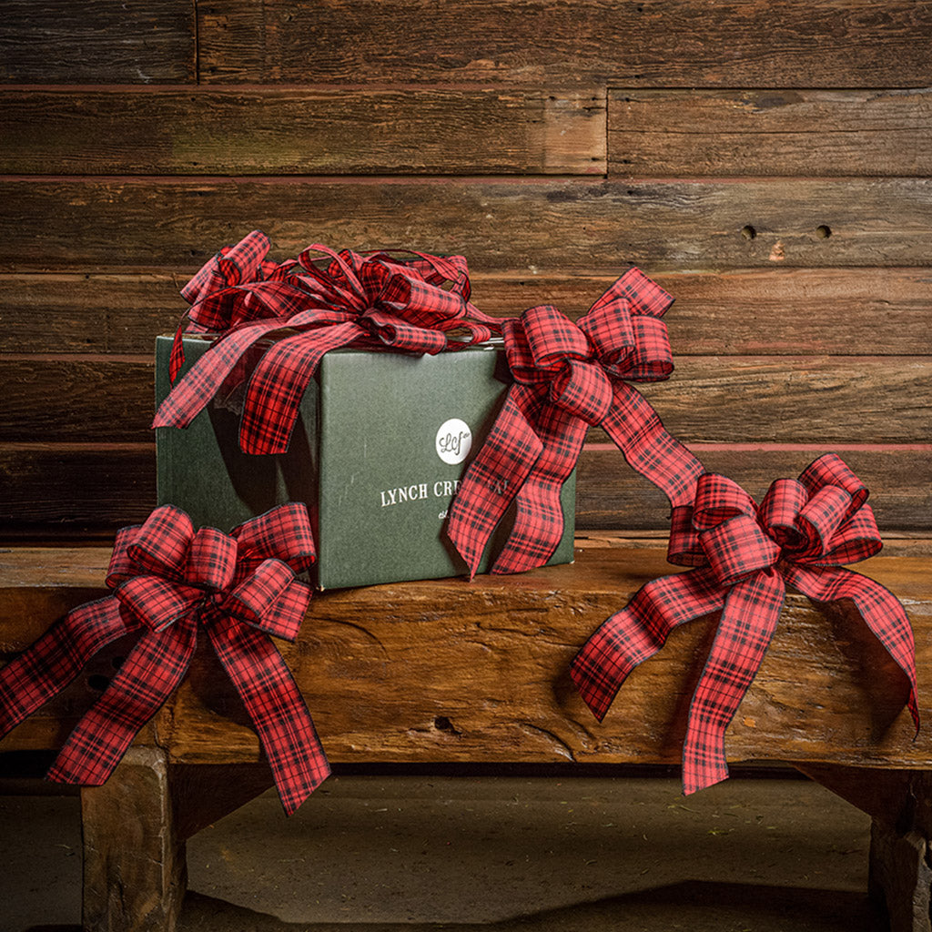 6 Red and Black plaid bows shown with a green shipping box sitting on a wood bench with a dark wood background.