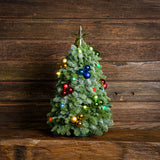Tabletop tree of noble fir with a string of battery-operated vintage-style colored lights, a gold star topper, and clusters of blue, red, green, and gold balls on a wooden table against a wood wall