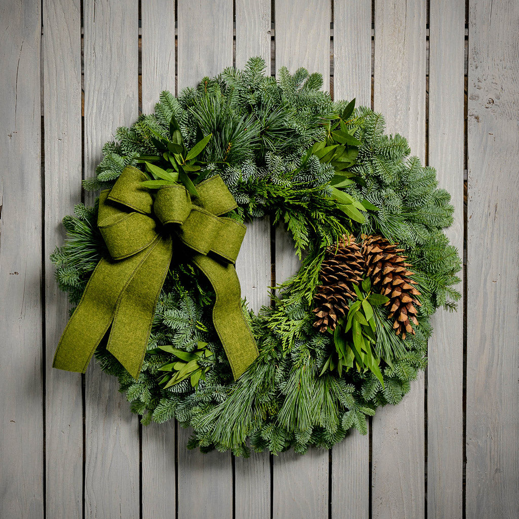 Christmas wreath with bay leaves, pine cones with a moss green brushed linen bow on a light wood background.