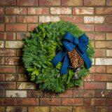 Holiday wreath made of noble fir, incense cedar, white pine, and California bay leaves with 6"- 8" white pine cones, burnished silver bells hanging from a jute string, and a navy blue brushed linen bow hanging on a brick wall