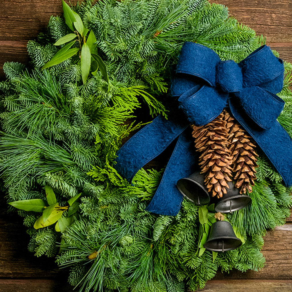 Holiday wreath made of noble fir, incense cedar, white pine, and California bay leaves with 6"- 8" white pine cones, burnished silver bells hanging from a jute string, and a navy blue brushed linen bow close up