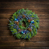 A holiday wreath of noble fir and western red cedar with a ring of faux metallic-blue berries and frosted branches, 3 blue and silver ball clusters, and 8 Australian cones on a wood background.