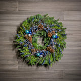 A holiday wreath of noble fir and western red cedar with a ring of faux metallic-blue berries and frosted branches, 3 blue and silver ball clusters, and 8 Australian cones on a wood background.