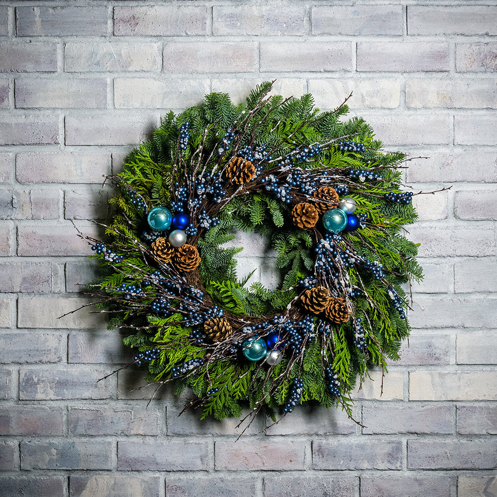 A holiday wreath of noble fir and western red cedar with a ring of faux metallic-blue berries and frosted branches, 3 blue and silver ball clusters, and 8 Australian cones on a white brick background.
