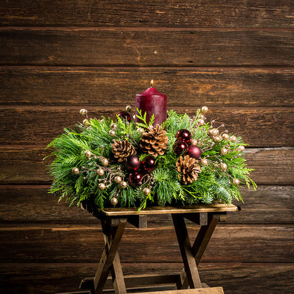 Centerpiece made of  verdant fresh noble fir, white pine, and incense cedar decorated with glittery beads and shiny burgundy balls, natural pinecones and a deep burgundy pillar candle on a small wood table.