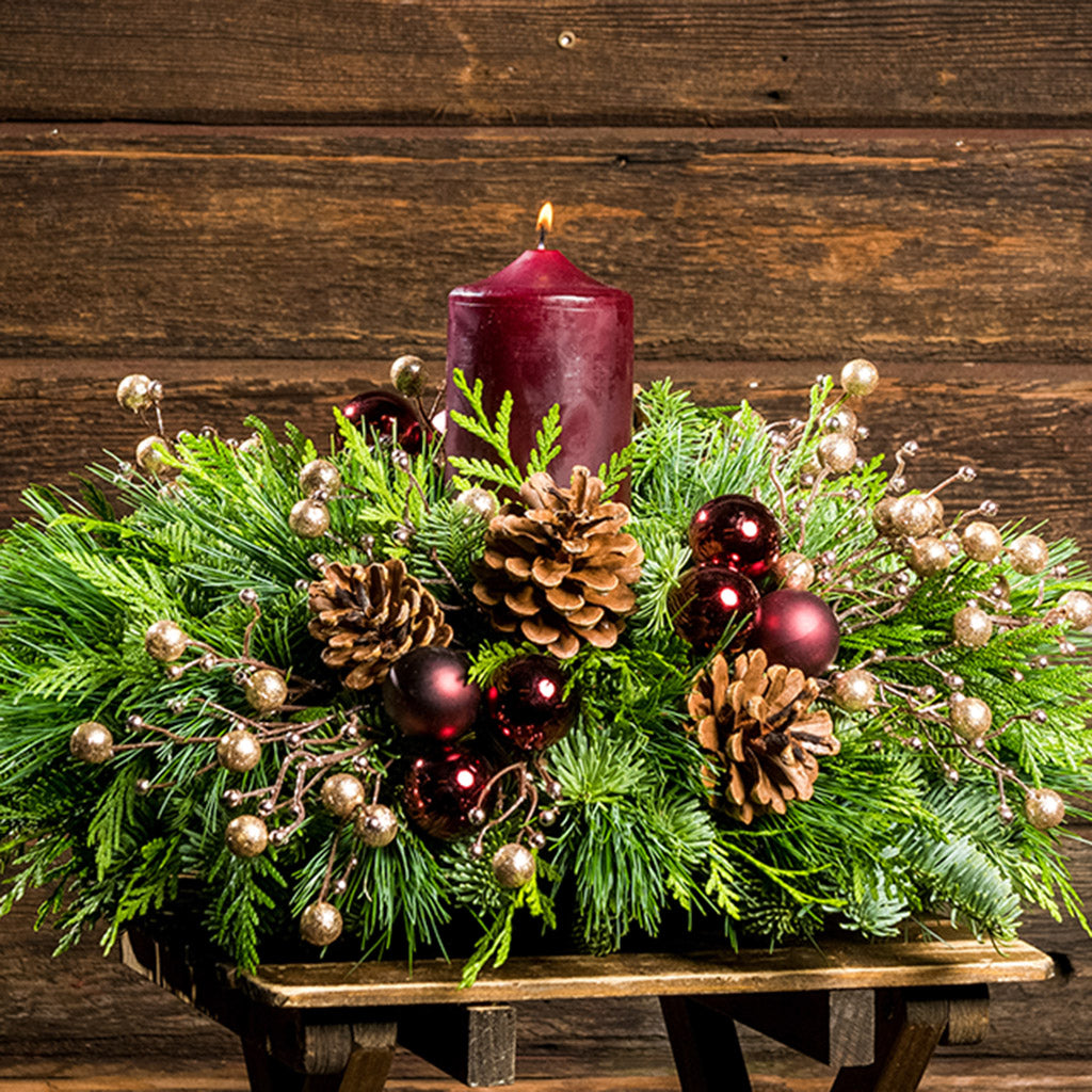 Centerpiece made of  verdant fresh noble fir, white pine, and incense cedar decorated with glittery beads and shiny burgundy balls, natural pinecones and a deep burgundy pillar candle on a small wood table.