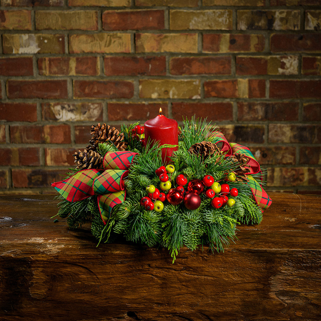 Centerpiece made of noble fir, cedar, and pine with ponderosa pine cones, Australian pine cones, red ball clusters, red and green faux berry picks, red-and-green plaid bow tucks, and a red pillar candle displayed on a wooden table against a brick wall