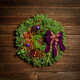 Holiday wreath made of noble fir, incense cedar, white pine, and bay leaf with purple and copper balls, gold pinecones and leaves, branches of multi-colored berries, and a purple velvet and shimmery copper double bow on a dark wood background.