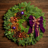 Holiday wreath made of noble fir, incense cedar, white pine, and bay leaf with purple and copper balls, gold pinecones and leaves, branches of multi-colored berries, and a purple velvet and shimmery copper double bow on a dark wood background.