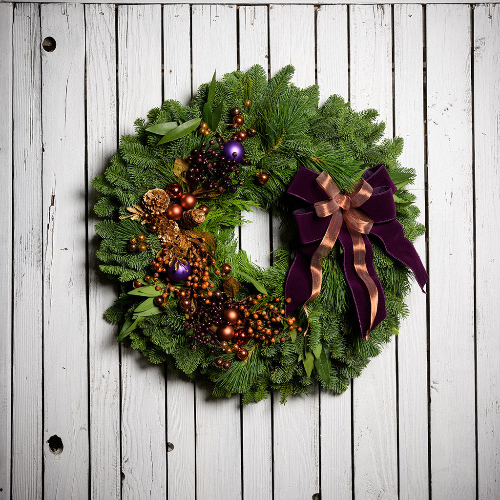 Holiday wreath made of noble fir, incense cedar, white pine, and bay leaf with purple and copper balls, gold pinecones and leaves, branches of multi-colored berries, and a purple velvet and shimmery copper double bow on a white wood background.