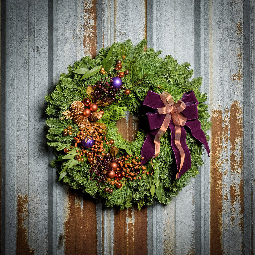 Holiday wreath made of noble fir, incense cedar, white pine, and bay leaf with purple and copper balls, gold pinecones and leaves, branches of multi-colored berries, and a purple velvet and shimmery copper double bow on a rustic metal background.