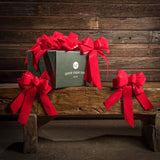 6 Bright Red bows shown with a green shipping box sitting on a wood bench with a dark wood background.