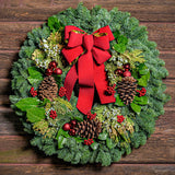 Holiday wreath made of noble fir, incense cedar, juniper, and salal leaves with ponderosa pine cones, faux red berry clusters, ball clusters, and a red bow on a wooden background
