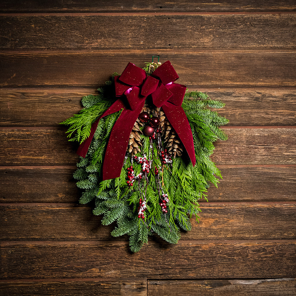 A swag made of noble pine and incense cedar with natural white-pine pinecones, shiny burgundy balls and glittery red berry branches with a sparkling velvet burgundy bow hanging on a wood wall.