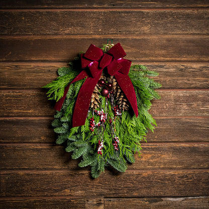 A swag made of noble pine and incense cedar with natural white-pine pinecones, shiny burgundy balls and glittery red berry branches with a sparkling velvet burgundy bow hanging on a wood wall.