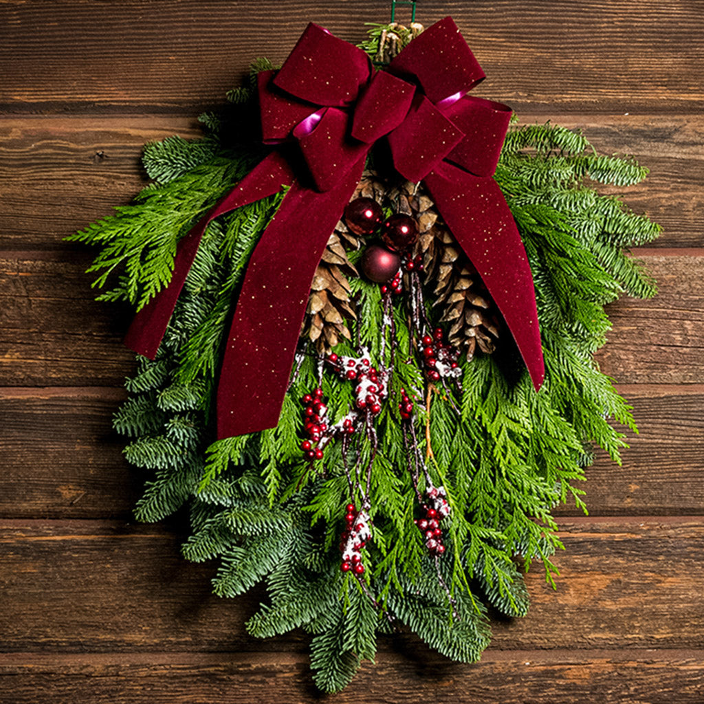 A swag made of noble pine and incense cedar with natural white-pine pinecones, shiny burgundy balls and glittery red berry branches with a sparkling velvet burgundy bow hanging on a wood wall.