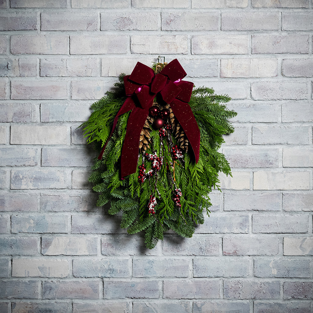A swag made of noble pine and incense cedar with natural white-pine pinecones, shiny burgundy balls and glittery red berry branches with a sparkling velvet burgundy bow hanging on a white brick wall.