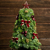 A small tree made of sprigs of fresh and fragrant noble fir evergreens, and decorated with burgundy bows with gold edges and shiny accents of burgundy balls and gold berries, topped with a burgundy star ornament with a dark wood background. 