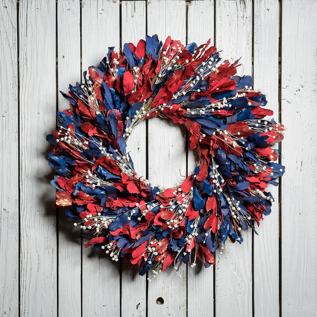 22" botanical wreath is handmade with red and blue integrifolia leaves and white flax on a white wood fence background.