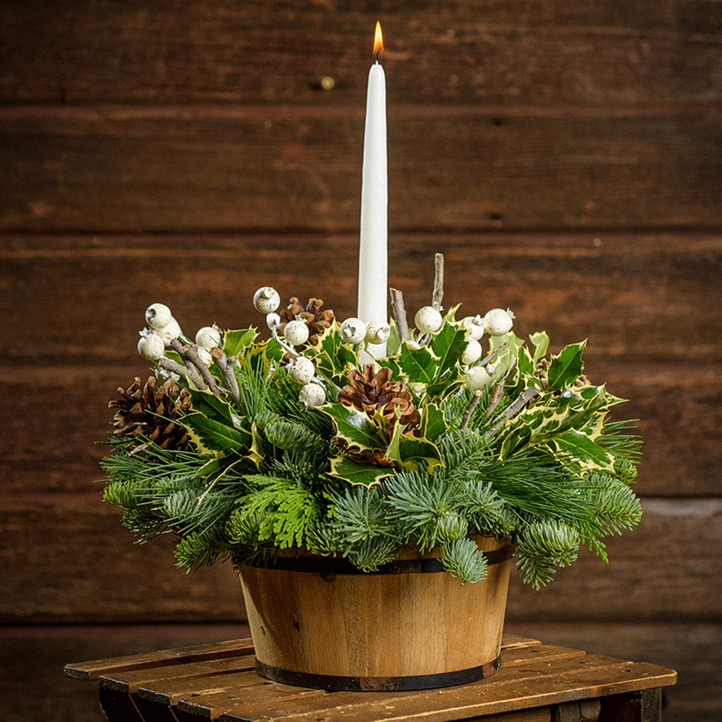 An arrangement made of noble fir, pine, and cedar with a wooden barrel, variegated holly, ponderosa pinecones, wooden sticks, white berry clusters, and an ivory taper candle with a dark wood background.