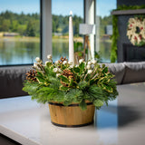 An arrangement made of noble fir, pine, and cedar with a wooden barrel, variegated holly, ponderosa pinecones, wooden sticks, white berry clusters, and an ivory taper candle sitting on a counter.
