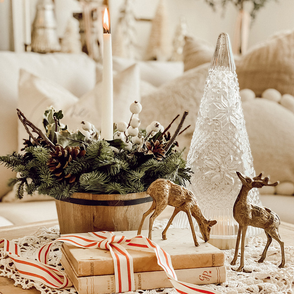An arrangement made of noble fir, pine, and cedar with a wooden barrel, variegated holly, ponderosa pinecones, wooden sticks, white berry clusters, and an ivory taper candle sitting on table.
