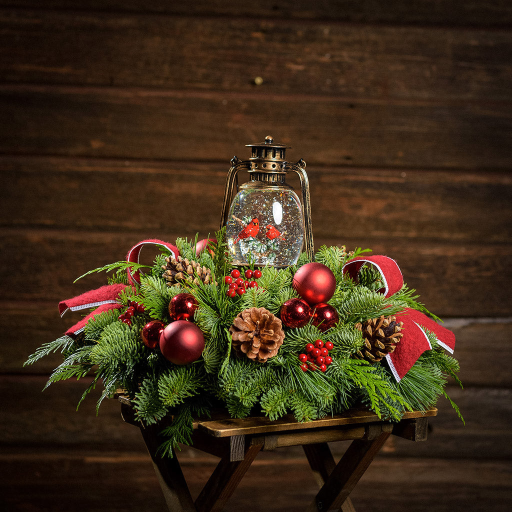 An arrangement of noble fir, incense cedar, and white pine with red ball clusters, pinecones, red berry clusters, red linen bows with white edges, and cardinals in a globe lantern with a wood background.